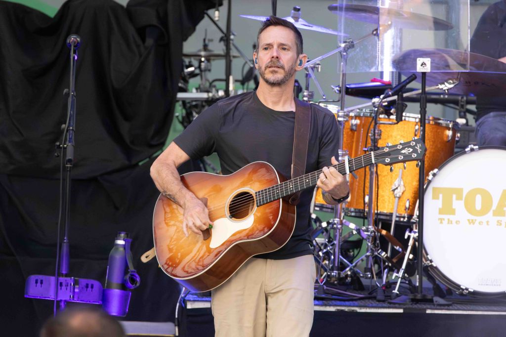 A musician playing an acoustic guitar on stage, wearing a black shirt and beige pants, with a drum set and microphone in the background.