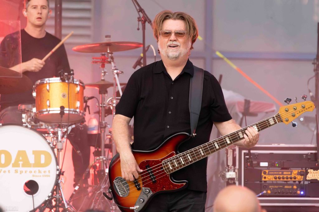 A musician playing a bass guitar on stage with a drummer in the background. The bassist is wearing sunglasses and a black shirt, smiling while performing.