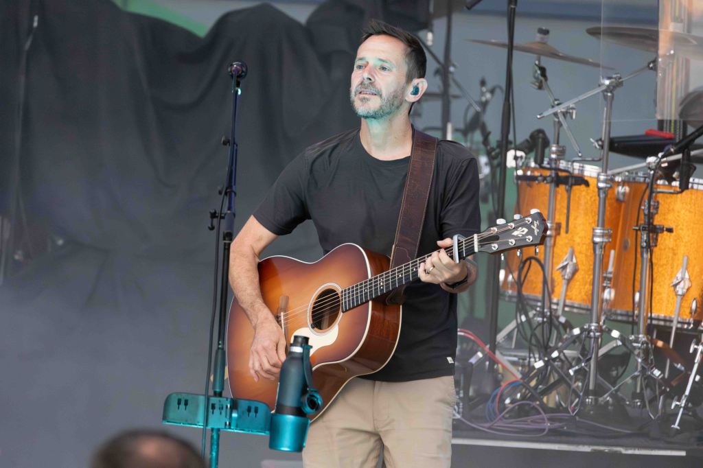 A musician performing on stage, playing an acoustic guitar and singing, with a blurred drum set and stage backdrop in the background.