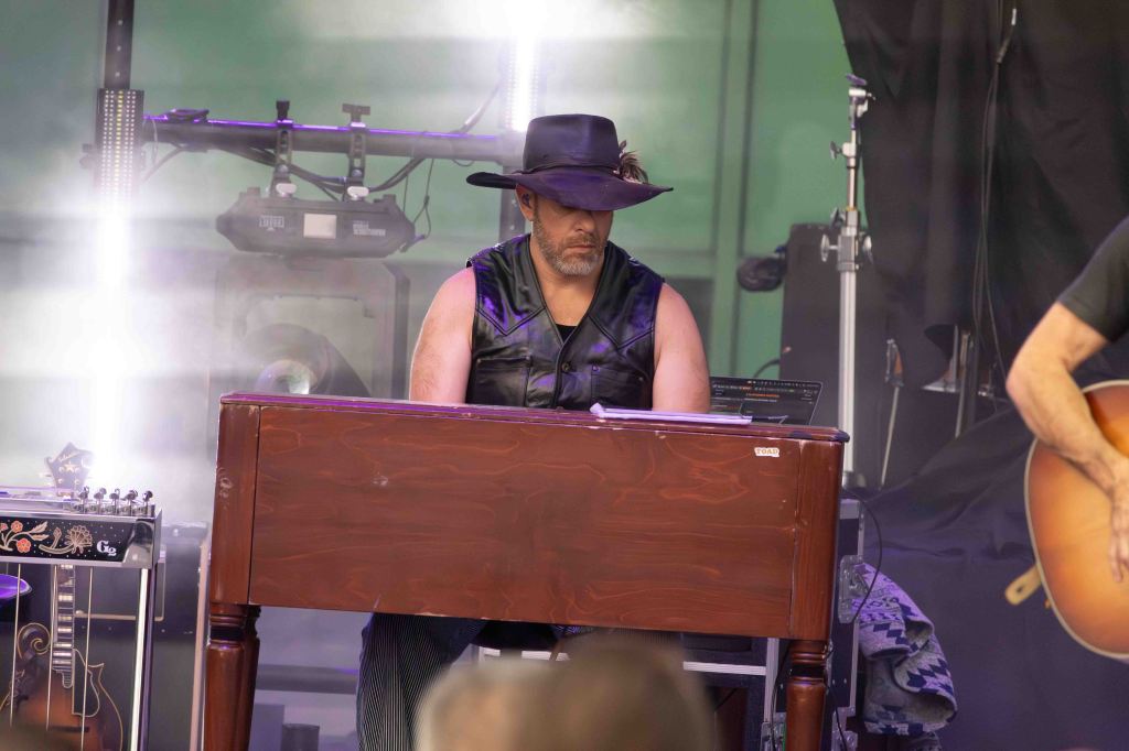 A musician playing a wooden keyboard on stage, wearing a black vest and hat, with lighting effects and instruments in the background.