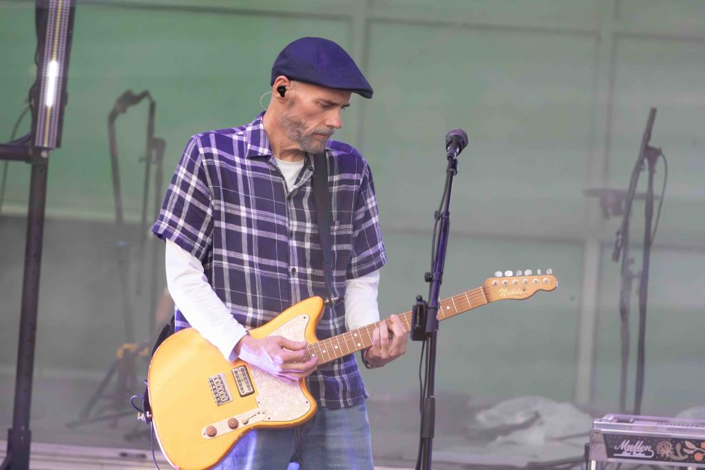 A musician wearing a plaid shirt and hat plays an electric guitar on stage, with microphones and equipment in the background.