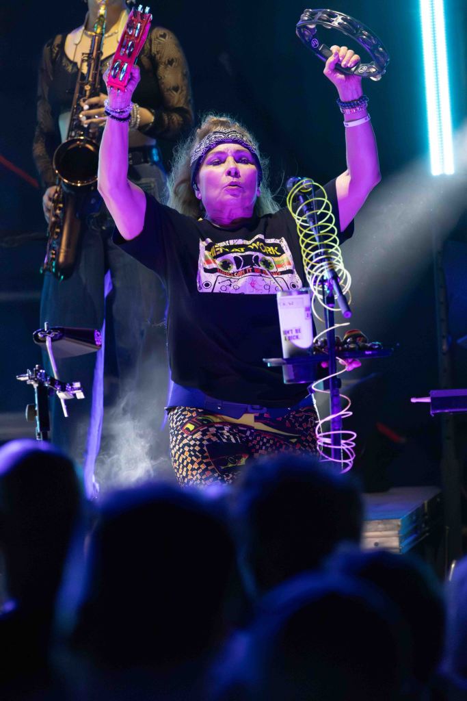 A female musician energetically performing on stage, holding a tambourine and exhibiting vibrant expressions. She is wearing a graphic t-shirt and colorful patterned pants, with a headband in her hair, surrounded by fog and stage lights.