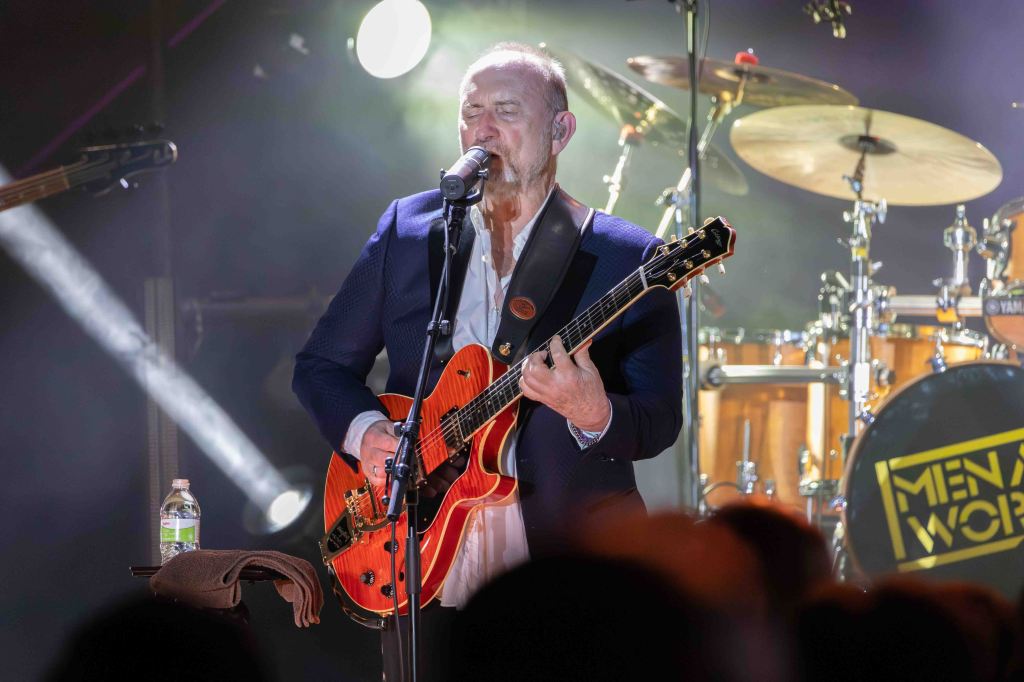 A musician performing on stage, singing into a microphone while playing an orange guitar. The background features drums and stage lights.