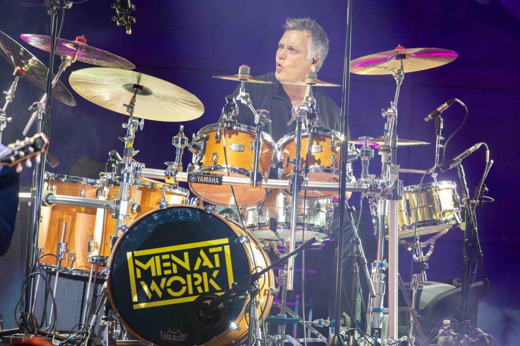 A drummer performing on stage with a drum set, featuring a bass drum displaying 'MEN AT WORK.' The drummer appears focused with various cymbals and microphones around him.