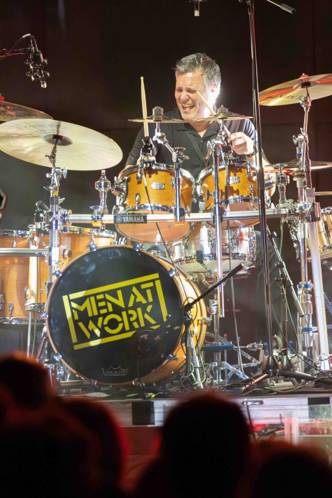A drummer performs on stage, smiling while playing a wooden drum set featuring a bass drum with the logo 'MEN AT WORK'.