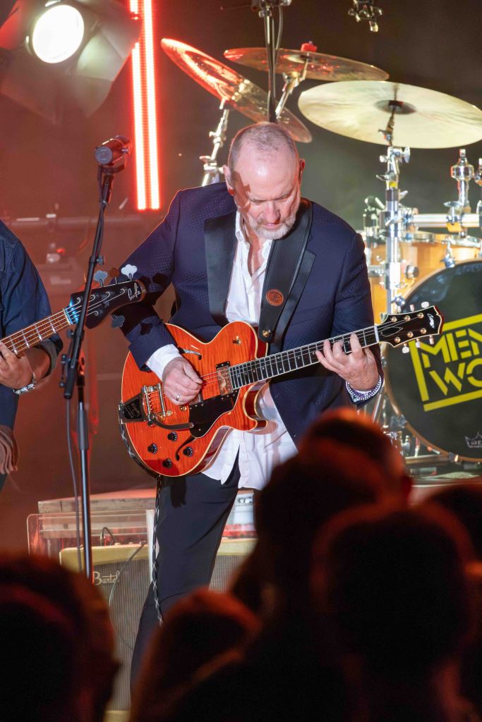 A musician in a black suit performs on stage, playing an orange electric guitar. Bright stage lights and a drum set are visible in the background, with an audience watching in the foreground.