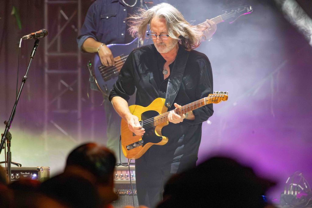 Musician playing electric guitar on stage with a band, surrounded by colorful stage lights and fog.