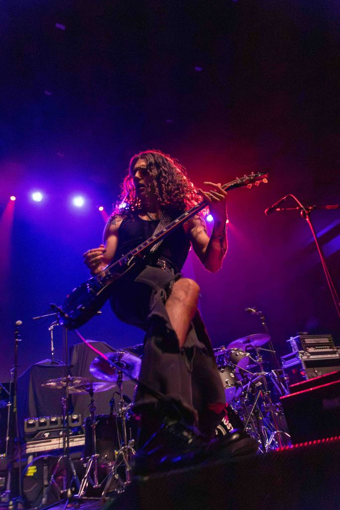 A musician playing an electric guitar on stage, with long curly hair and wearing a sleeveless top, illuminated by colorful stage lights.