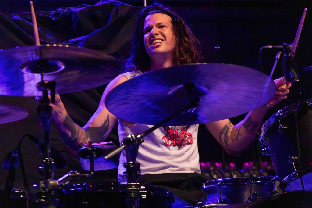 A drummer performing energetically on stage, wearing a sleeveless shirt and smiling while playing the drums.