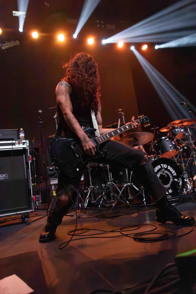 A musician performing on stage, passionately playing an electric guitar with long hair flowing, wearing a sleeveless black top and ripped black pants, with stage lights illuminating the scene.