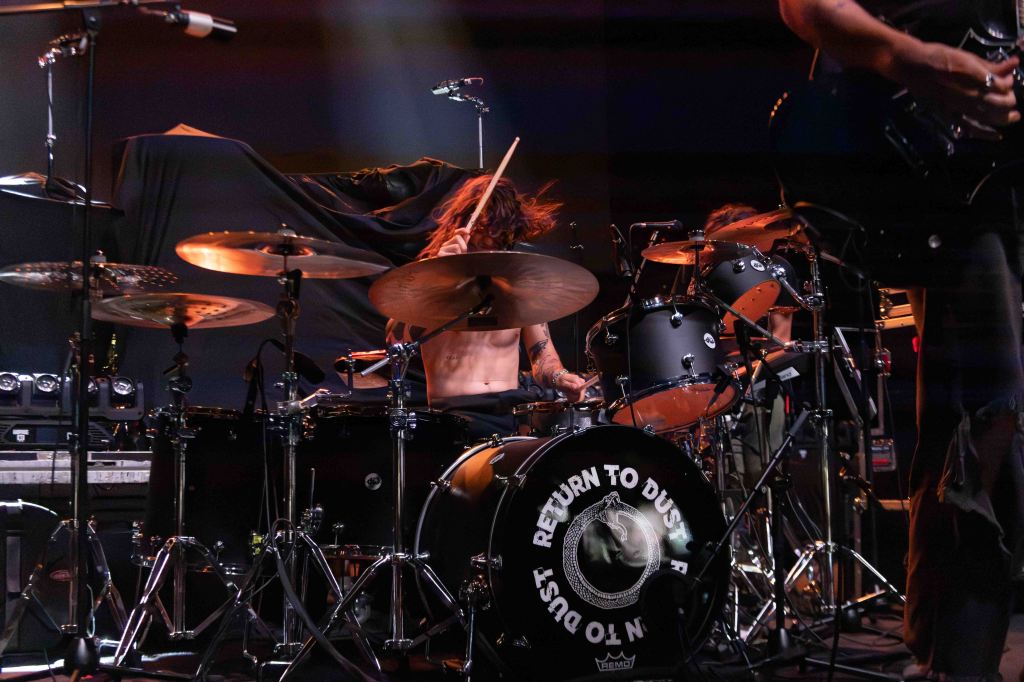 A drummer performing on stage in front of a dynamic backdrop, with a focus on the drum set featuring a logo that reads 'RETURN TO DUST'. The drummer is captured mid-performance, energetically playing the drums.