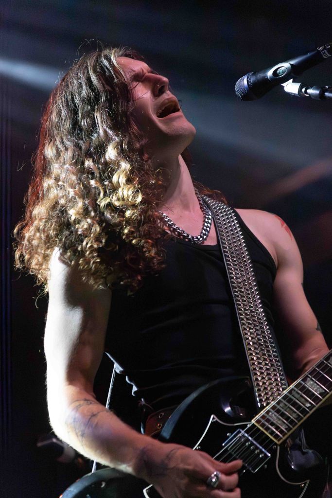 A musician passionately performing on stage, singing into a microphone while playing an electric guitar. He has long, curly hair and is wearing a sleeveless top.