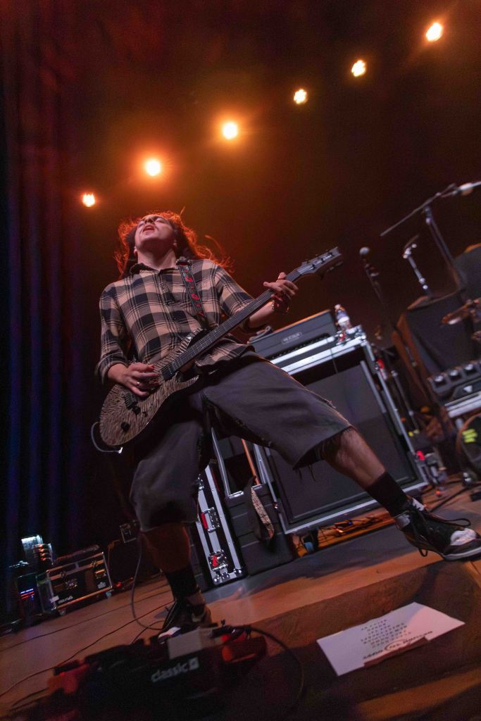 A musician passionately playing an electric guitar on stage, wearing a plaid shirt and shorts, with dynamic stage lighting in the background.