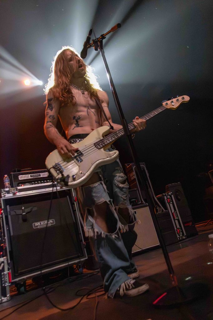 A shirtless musician with long hair playing an electric bass guitar on stage, illuminated by dramatic stage lights. He wears ripped jeans and has tattoos visible on his arms.