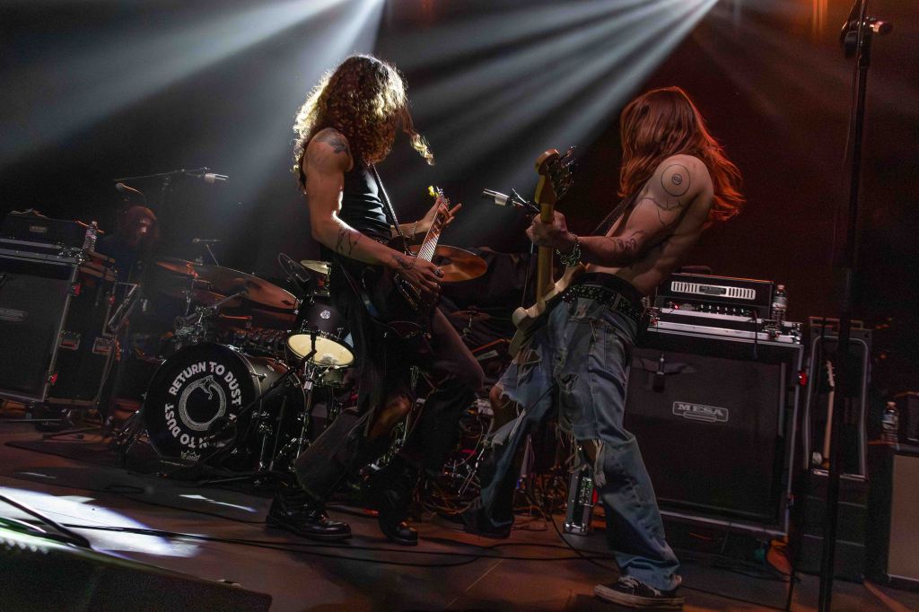 Two musicians performing passionately on stage, with one playing electric guitar and the other playing bass. The scene features dramatic lighting and a drum set in the background.