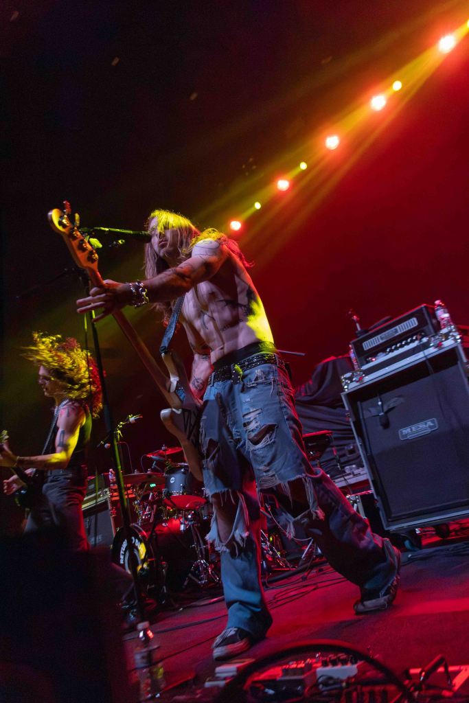 A shirtless musician playing a bass guitar on stage, surrounded by dramatic lighting. The performer has long hair and is wearing ripped jeans. The background features other band members and musical instruments.