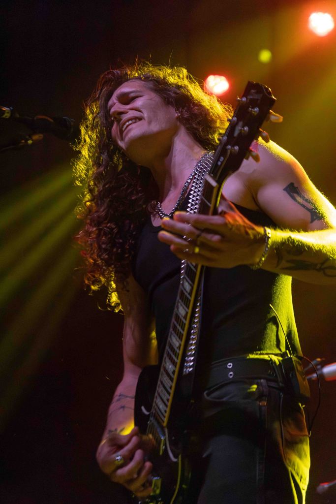 A musician passionately playing an electric guitar on stage, with curly hair and a black tank top, illuminated by stage lights.