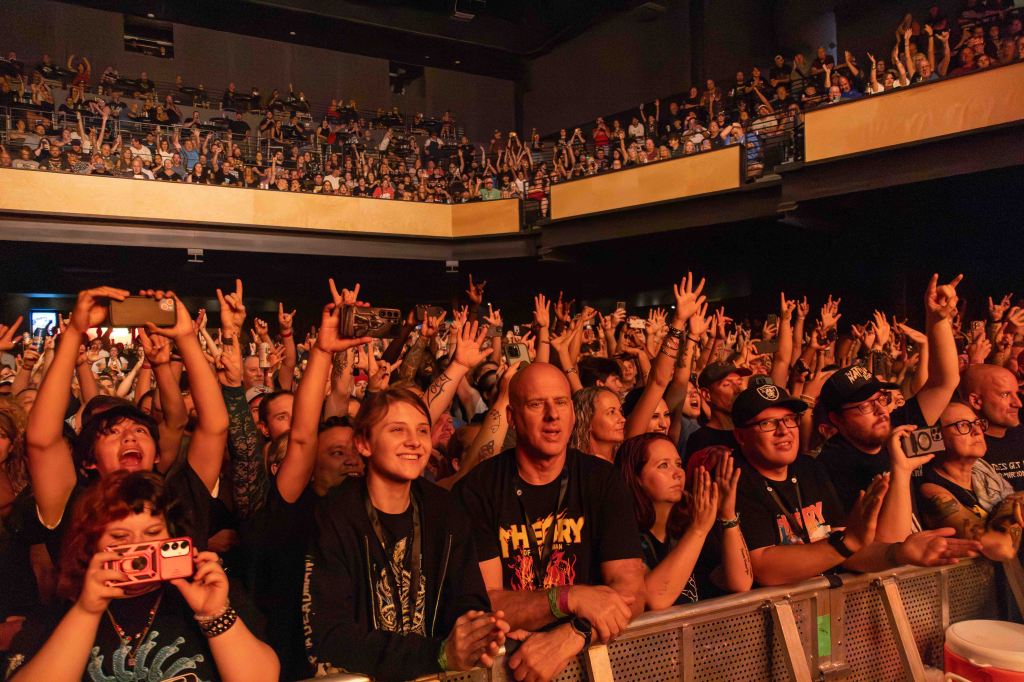 A large crowd at a concert with people raising their hands and cheering, showcasing a lively atmosphere. Many attendees are wearing band merchandise and some are taking photos.