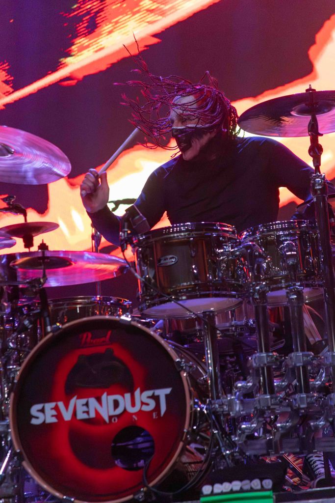 Drummer performing on stage with flying hair, wearing a black outfit, and playing a drum set with a logo of SEVENDUST prominently displayed on the bass drum.