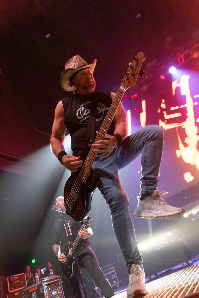 A musician passionately playing the bass guitar on stage, wearing a cowboy hat and a sleeveless shirt, with dramatic stage lighting in the background.