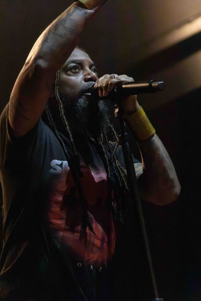 A close-up of a performer with long dreadlocks, passionately singing into a microphone while raising one hand. They are wearing a black shirt featuring a colorful graphic design. The background is dimly lit, creating a vibrant atmosphere.