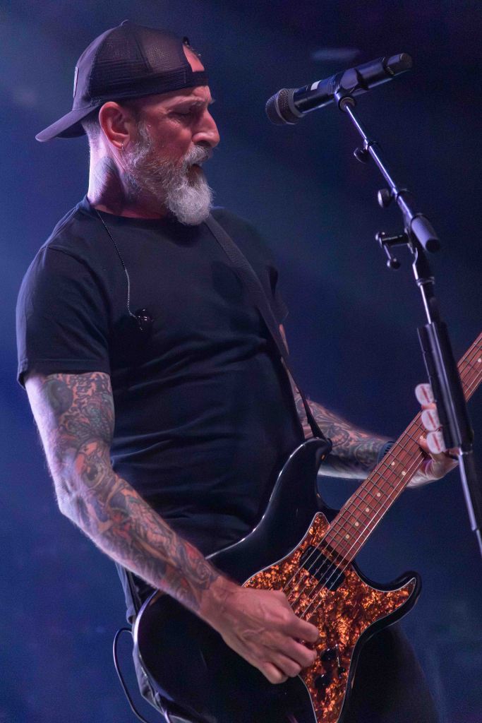 A musician with a beard and tattoos playing an electric guitar on stage, wearing a black t-shirt and a cap. The image captures the intensity of the performance with colorful stage lighting.