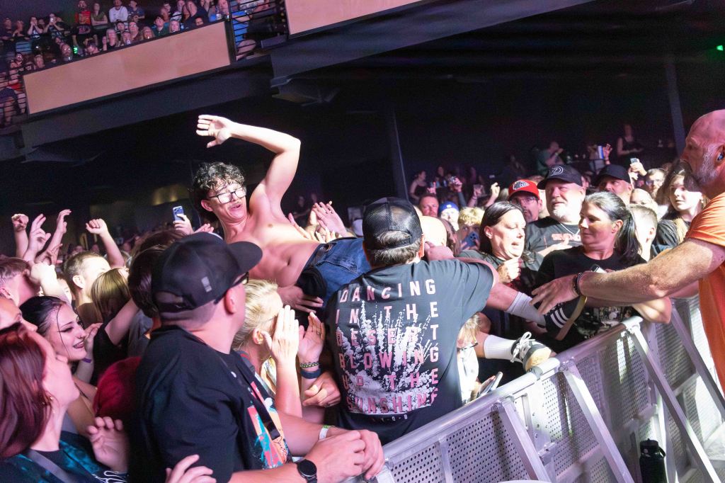 A lively concert scene with an energetic crowd. A shirtless male is crowd surfing while surrounded by enthusiastic fans, some reaching out to catch him. The atmosphere is filled with excitement and engagement.