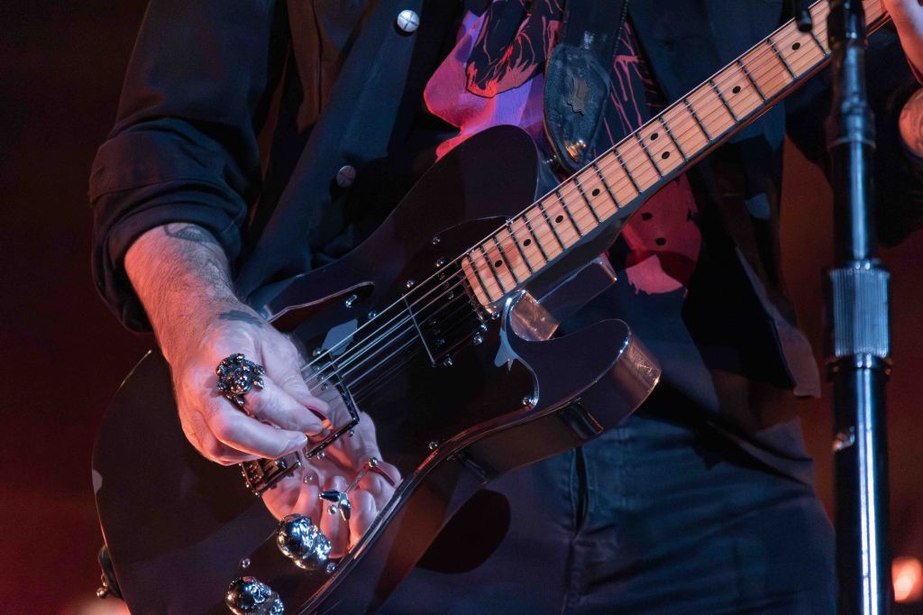 Close-up of a musician's hand playing a black electric guitar, adorned with striking rings, wearing a graphic t-shirt and a dark jacket.