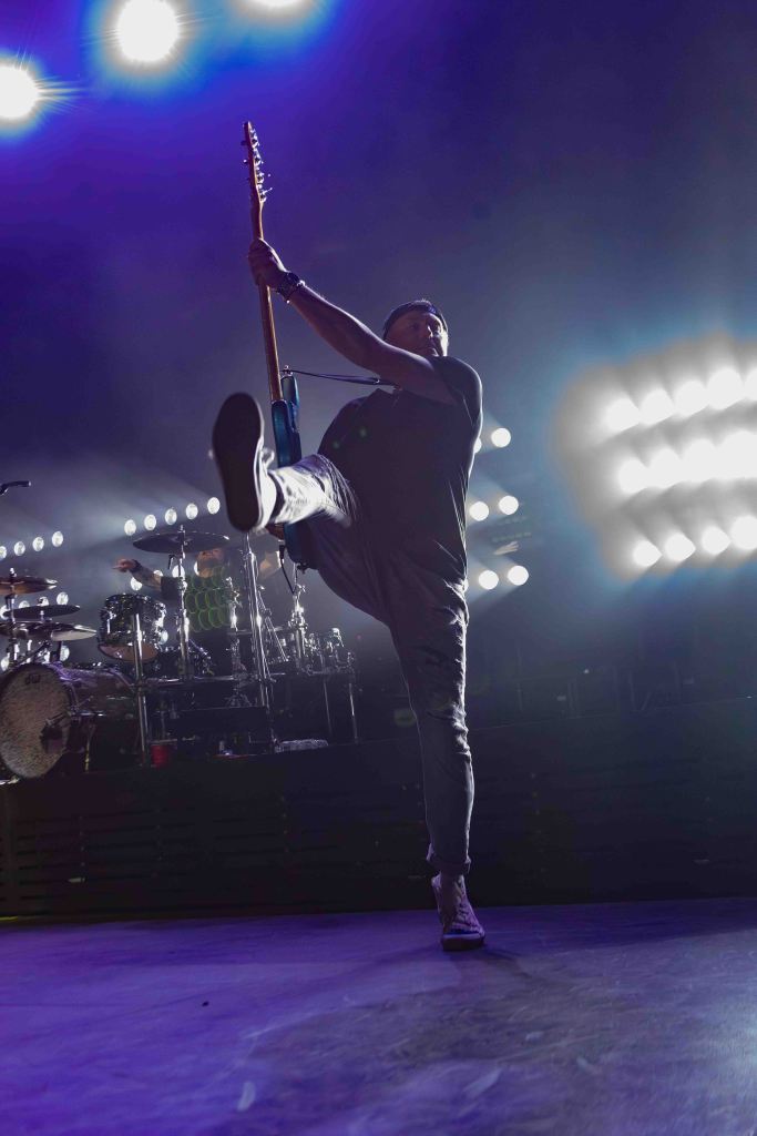 A musician energetically performing on stage with one leg raised and holding a guitar, illuminated by bright stage lights.