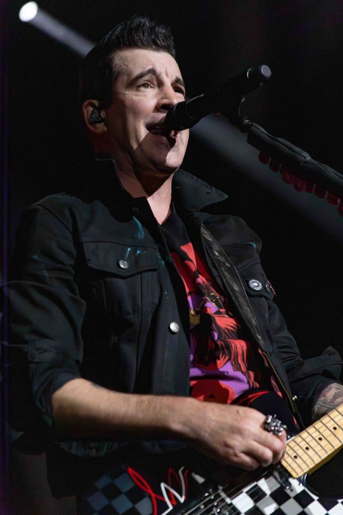 A male musician singing into a microphone while holding an electric guitar. He is wearing a black jacket over a colorful T-shirt and appears to be performing on stage.