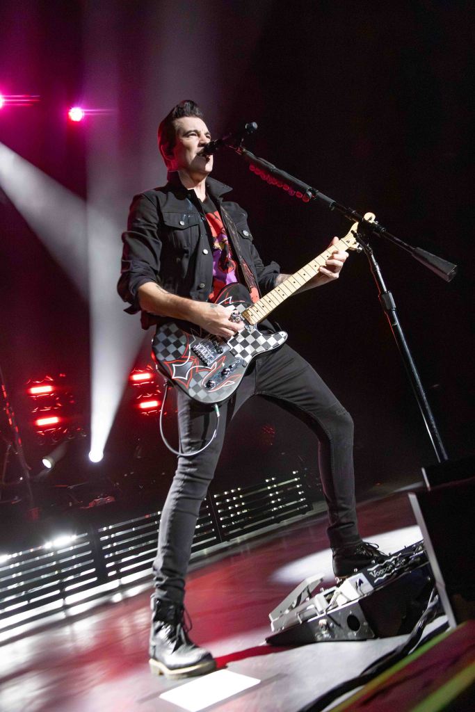 A male guitarist performing on stage with a colorful electric guitar, wearing a black jacket and a graphic t-shirt, under bright stage lights.