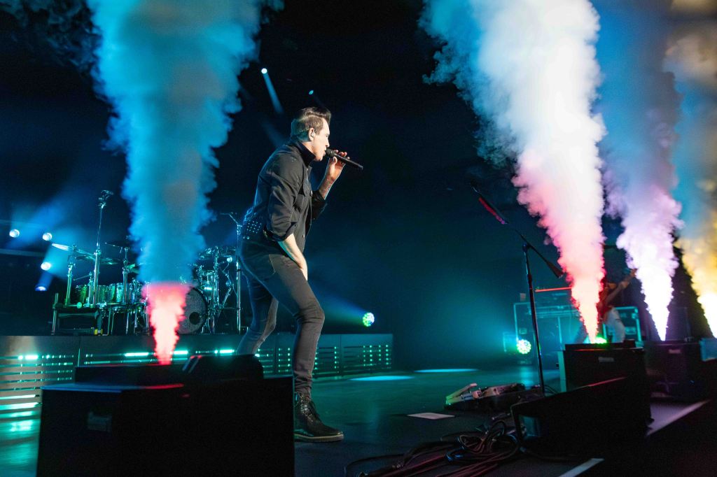 A singer performing on stage with colorful smoke effects and dramatic lighting, accompanied by a drum set in the background.