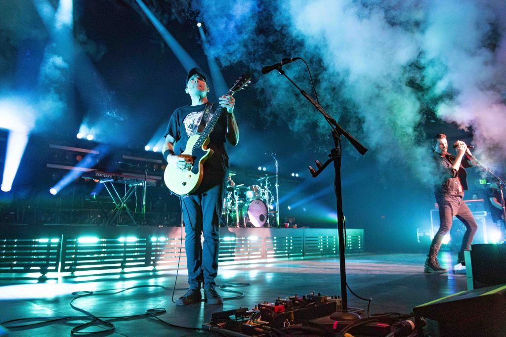 A guitarist playing an electric guitar on stage with colored lights and smoke effects in the background, accompanied by a vocalist and a drummer.