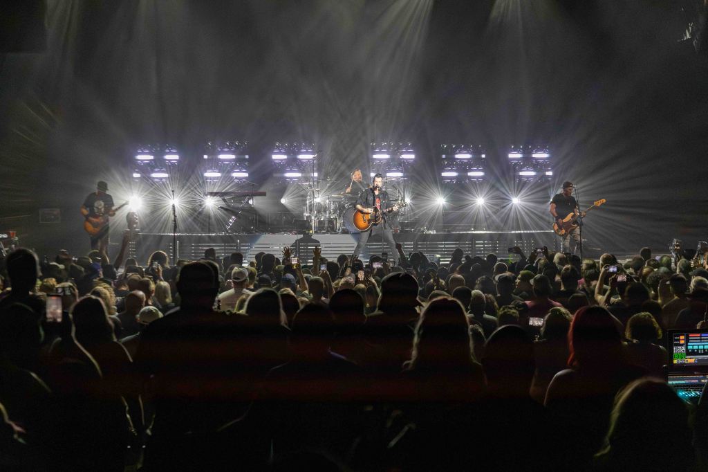 A live concert scene featuring a band performing on stage with bright lights and a captivated audience. The frontman is playing an acoustic guitar, flanked by band members on guitar, bass, and keyboards, while the crowd is holding up their phones to capture the performance.