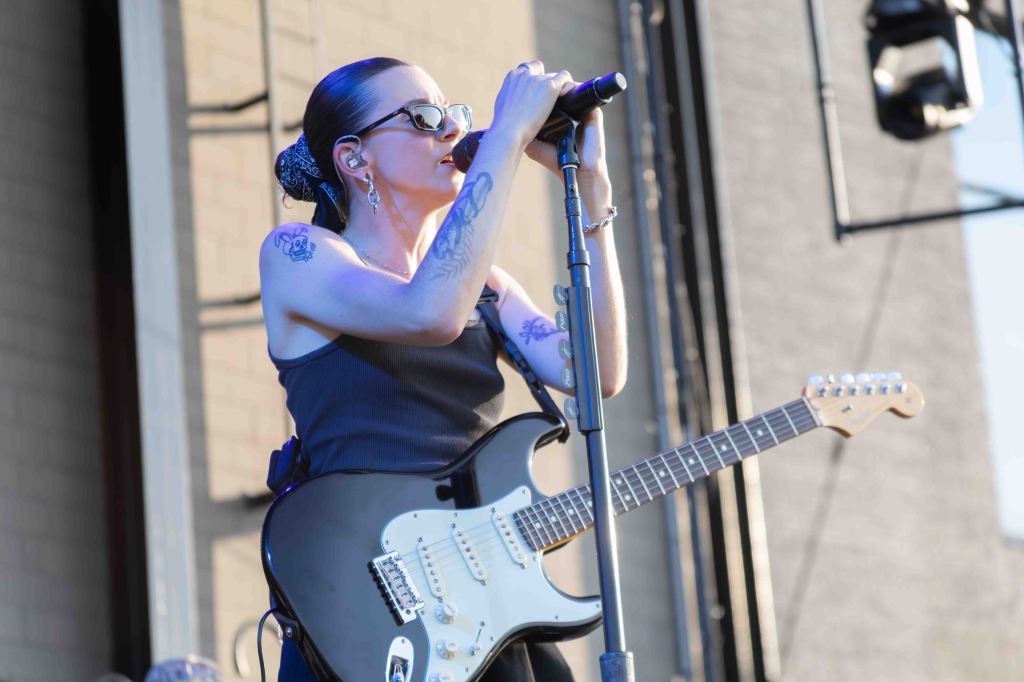 A female musician performing live on stage, playing an electric guitar and singing into a microphone, wearing sunglasses and a black tank top, with tattoos visible on her arms.