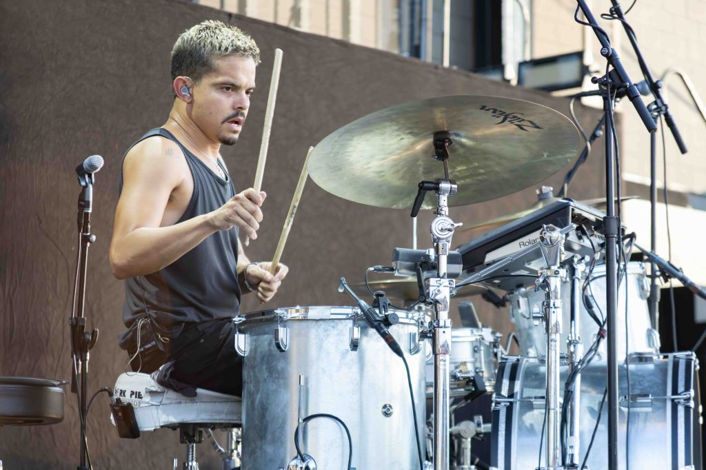 A drummer performing on stage, playing a drum set with a microphone nearby. The drummer is wearing a sleeveless shirt and focused on the music.