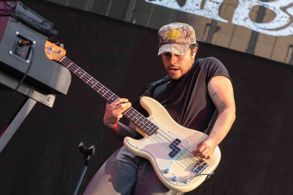 A musician playing a white bass guitar on stage, wearing a camouflage cap and a black t-shirt.