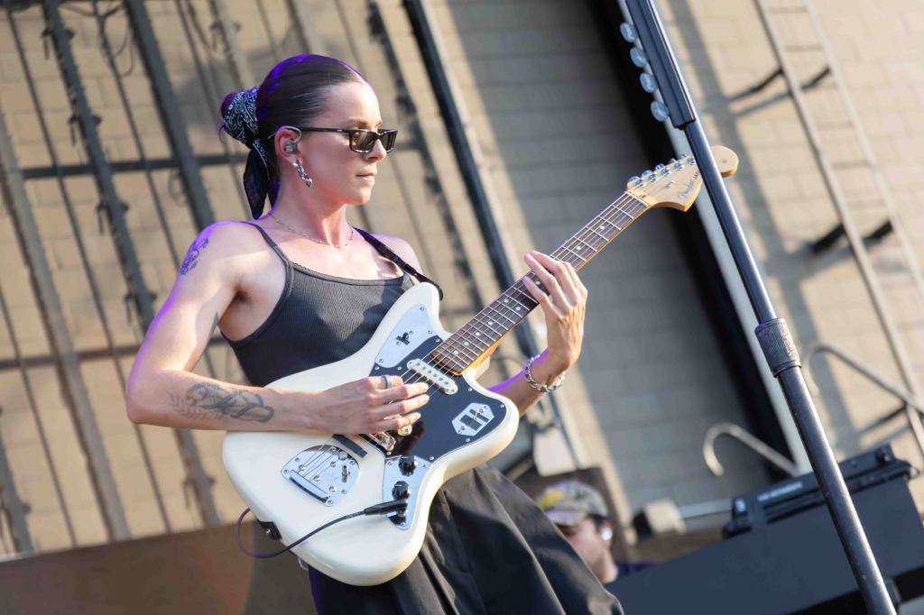 A woman wearing sunglasses and a black tank top plays an electric guitar on stage, with a microphone stand in the foreground and blurred audience members in the background.