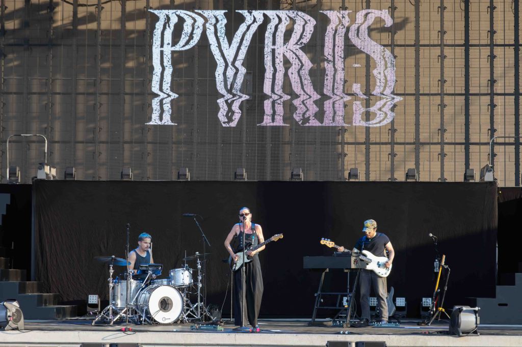 A band performing live on stage with the name 'PYRIS' displayed in large letters behind them. The group consists of three musicians: a female vocalist playing guitar, a drummer, and a male musician on keyboard and bass guitar.