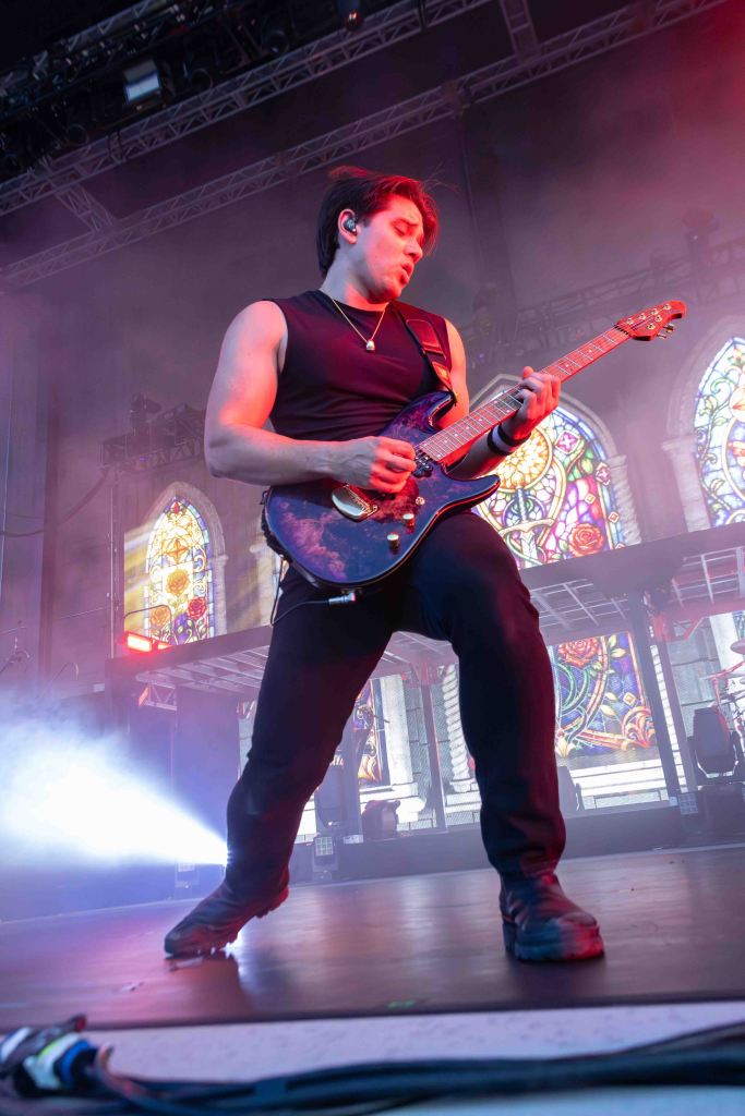 A male guitarist passionately performing on stage, illuminated by colorful lights, with stained glass windows in the background.