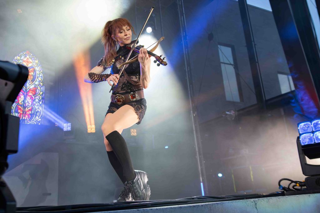 A female violinist performing on stage, wearing a stylish outfit, with dramatic lighting and a stained glass backdrop.