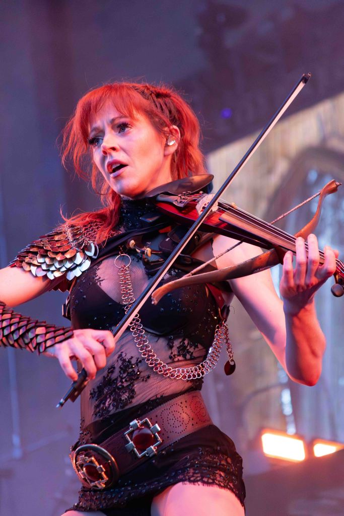 A female musician passionately performing onstage with a violin, wearing a striking costume featuring intricate details, leather straps, and a decorative belt.