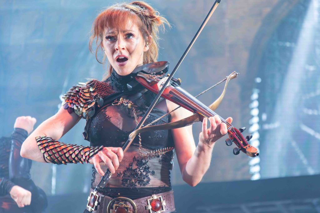 A spirited female musician playing a violin, dressed in an elaborate costume featuring armor-like elements, performing on stage with lights in the background.