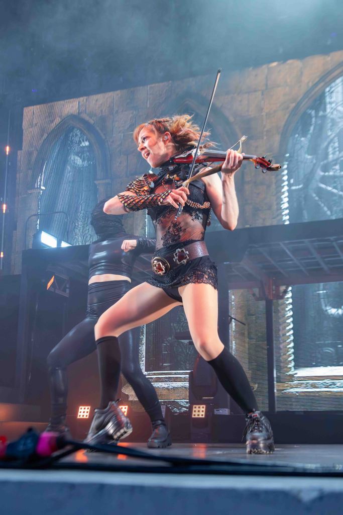 A female violinist performs energetically on stage, wearing a striking outfit and smiling. A dancer is in the background, and dramatic lighting enhances the concert atmosphere.