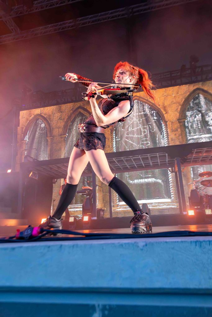A female musician performing on stage, playing the violin. She has bright red hair and is wearing a black outfit with thigh-high socks. The background features dramatic lighting and architectural elements.
