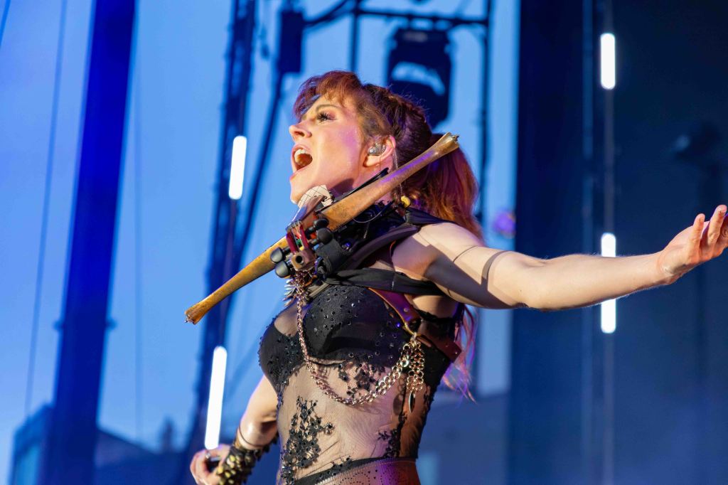 A female musician performing passionately on stage, holding a violin and singing, dressed in a black outfit with intricate details, illuminated by stage lights.