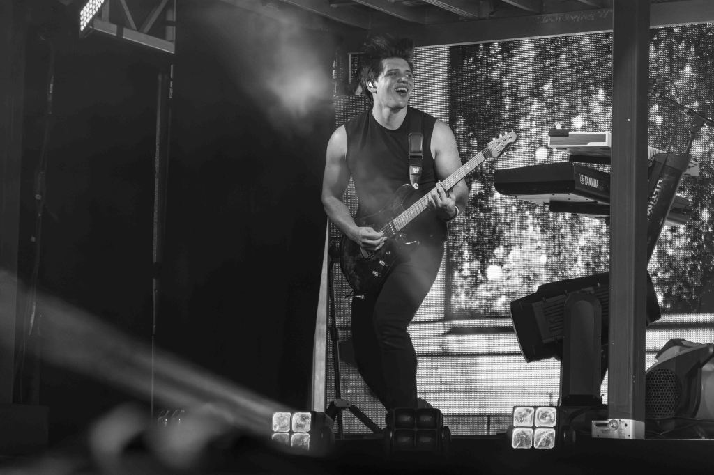 A guitarist energetically performing on stage with a keyboard setup in the background, captured in black and white.