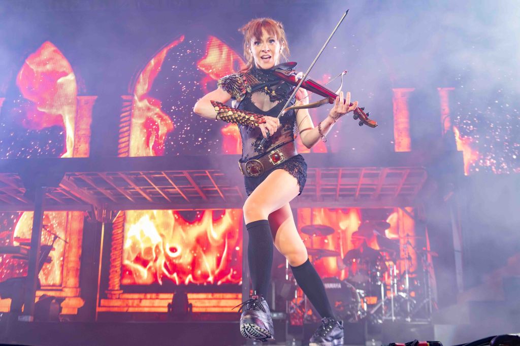 A female musician performing on stage, playing the violin while wearing a stylish outfit. The background features vibrant flames and dramatic lighting effects.