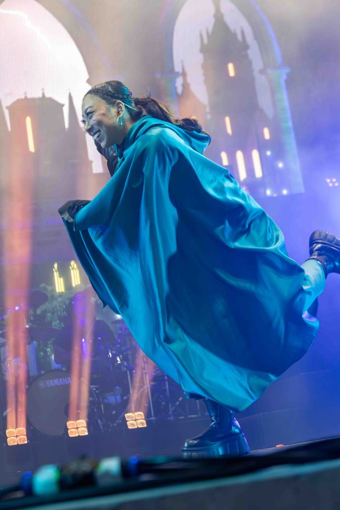 A performer wearing a bright blue cape smiles while dancing on stage, with colorful lights and a band setup in the background.