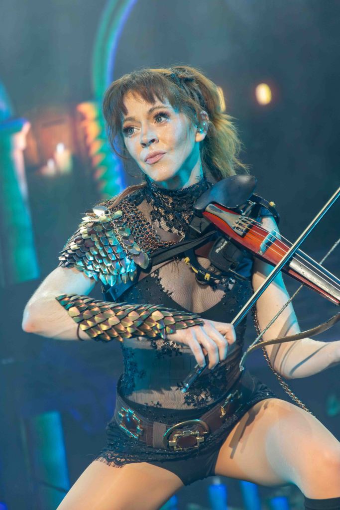 A female performer playing a violin on stage, wearing a unique costume featuring metallic accents and sheer fabric, with dramatic stage lighting in the background.